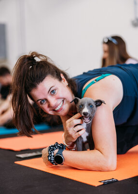 Yoga class attendant poses and smiles into the camera while puppy bites her finger