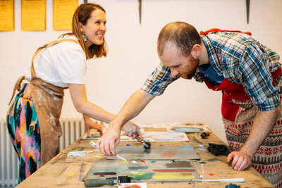 Two print workshop participants roll out paint ready to use to make Christmas card and gift wrap, both wearing Christmas aprons