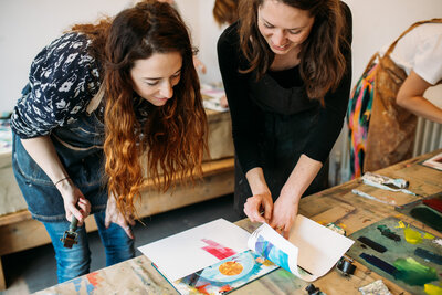 Two women, host and participant, stand by table and observe art being formed in front of their eyes as one of them peels of stencil and reveals print of birds as the fly into the sunset with landscape around, art supplies spread around the print