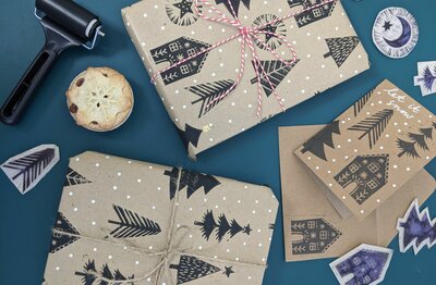 Christmas cards and presents wrapped in printed gift wrapped laid out on table together with printing tools and mince pie