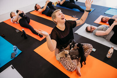 Excited looking yoga class attendant sits on yoga matt with lap covered with adorable puppies while other people stretch