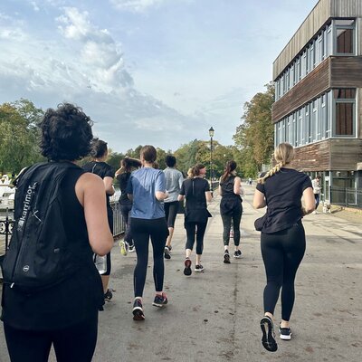 A group of people jogs together on a sunny day along a riverside path, bordered by trees and a modern building. The mood is active and communal.