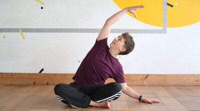 A person in a purple shirt and black pants performs a seated side stretch on a wooden floor. The wall behind is white with yellow and black accents.