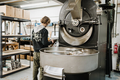 A person operates a large coffee roasting machine in an industrial setting. Shelves with boxes surround the workspace, creating a busy, focused atmosphere.