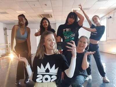 A group of six women pose joyfully in a dance studio, smiling and gesturing playfully. The room is bright, with casual attire and an upbeat atmosphere.