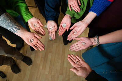 A group of seven people stand in a circle on a wooden floor, each extending a hand holding a ring. The mood is collaborative and inclusive.