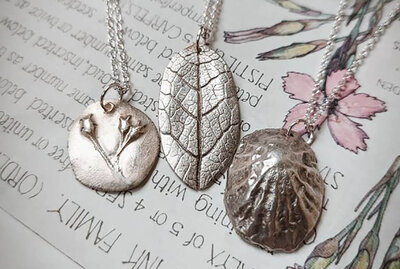 Three silver necklaces with nature-inspired pendants: a circular one with flower buds, a leaf-shaped design, and a textured oval shell. They rest on a botanical illustration and handwritten text, creating a serene, artistic mood.