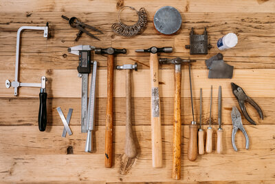 A neatly arranged set of jewelry-making tools on a wooden surface, including saws, hammers, calipers, files, and pliers, conveying precision and craftsmanship.