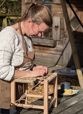 A woman in a white sweater and brown apron weaves a seat on a wooden stool outside a rustic cabin. The scene is sunny and conveys focus and craftsmanship.