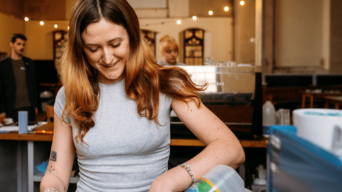 A woman stands with a cosy cafe scene behind. She is smiling as she pours milk