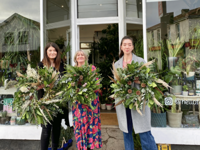 Three smiling people proudly showing off their lush, handmade wreaths outside a charming plant shop.
