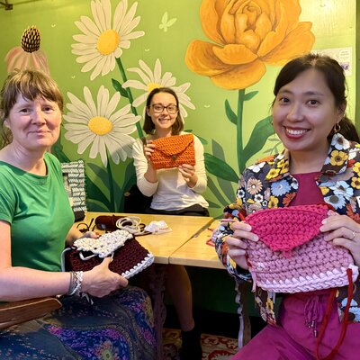 Three friends delight in crochet creations, against a cheerful floral backdrop.