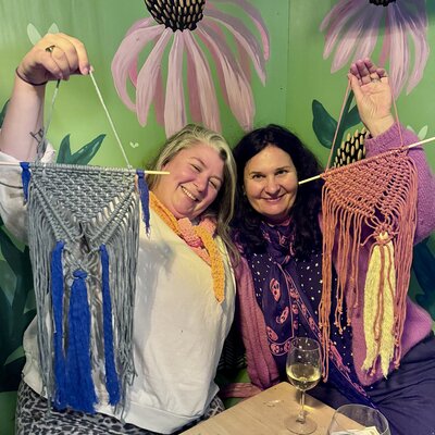 Joyful duo showing off their fabulous macramé creations in a cosy, creative setting.