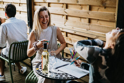 Two friends enjoying a playful art session with wine and laughter.