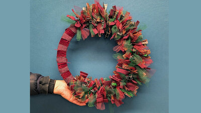 Hand holding a festive wreath of red and green ribbons against a blue wall.